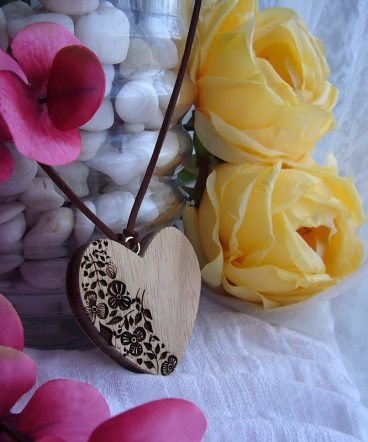 colgante corazón de madera grabado con flores y hojas sobre fondo blanco con flores rosas y amarillas a los lados