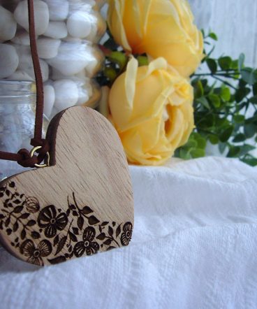 corazón de madera natural con un grabado de hojas y flores en la mitad izquierda, sobre fondo blanco y rosas amarillas al fondo