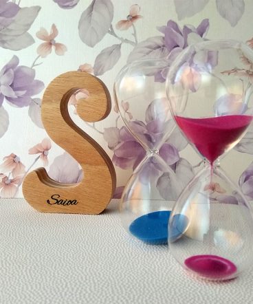 Letra S de madera personalizada con el nombre de Saioa sobre fondo con flores malva y dos relojes de arena a la derecha