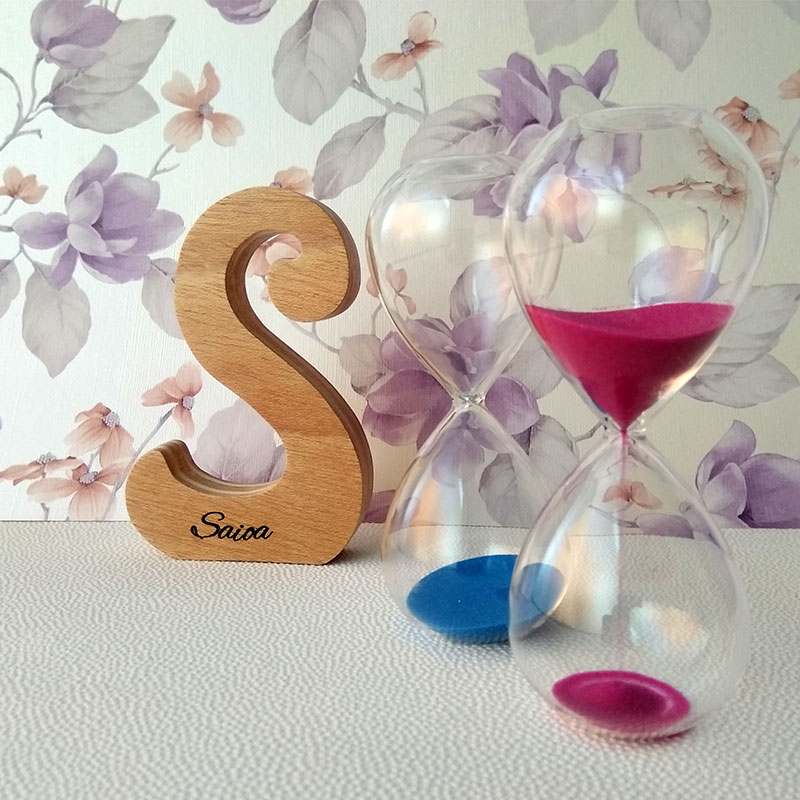 Letra S de madera personalizada con el nombre de Saioa sobre fondo con flores malva y dos relojes de arena a la derecha