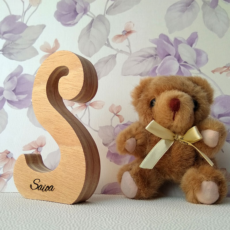 Letra S de madera personalizada con el nombre de Saioa sobre fondo con flores malva y osito de peluche a la derecha