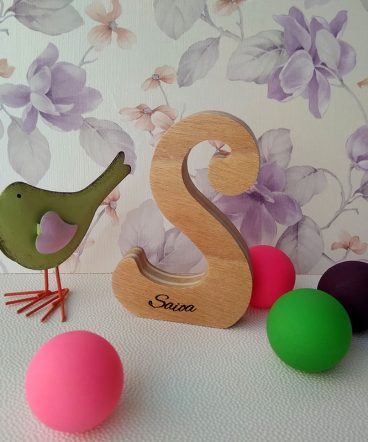 Letra S de madera personalizada con el nombre de Saioa sobre fondo con flores malva, pajarito ver y pelotas pequeñas de colores en la superficie
