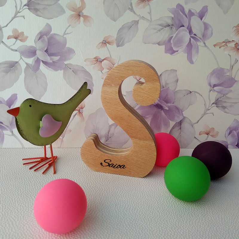 Letra S de madera personalizada con el nombre de Saioa sobre fondo con flores malva, pajarito ver y pelotas pequeñas de colores en la superficie