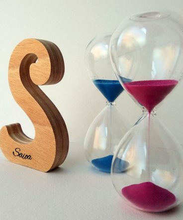 Letra S de madera personalizada con el nombre de Saioa sobre fondo con flores malva y dos relojes de arena a la derecha