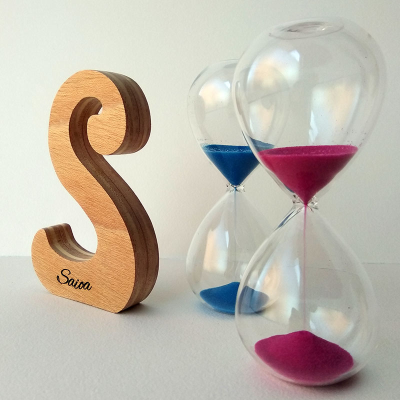 Letra S de madera personalizada con el nombre de Saioa sobre fondo con flores malva y dos relojes de arena a la derecha