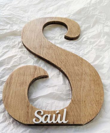 letra s de madera natural sobre papel arrugado blanco, decorada con el nombre de Saúl en blanco y en relieve pegado en la parte de abajo