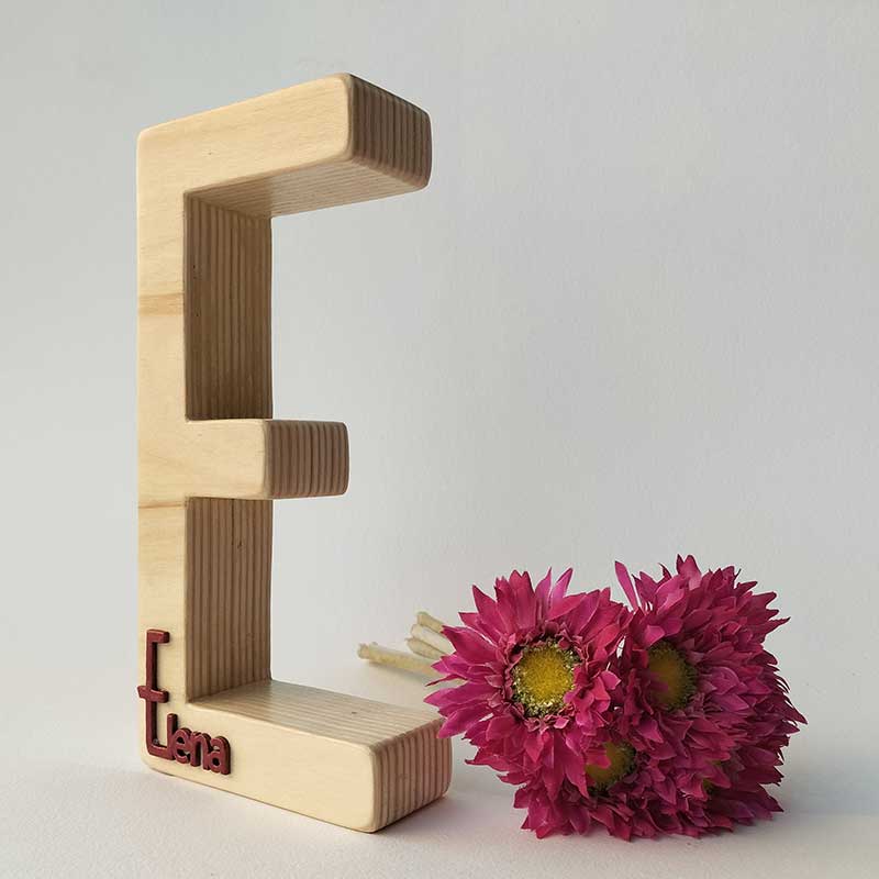 fondo blanco con letra e de madera maciza con el nombre de Elena en rojo en relieve pegado en la parte de abajo y flores fucsia a la derecha
