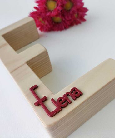 fondo blanco con letra e de madera maciza con el nombre de Elena en rojo en relieve pegado en la parte de abajo y flores fucsia al fondo