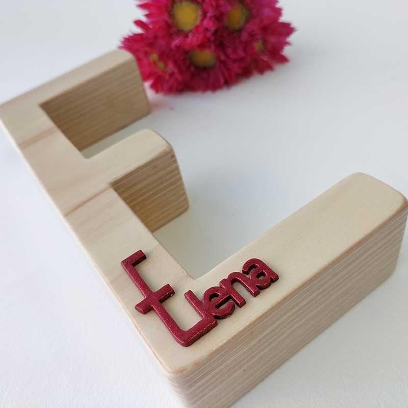 fondo blanco con letra e de madera maciza con el nombre de Elena en rojo en relieve pegado en la parte de abajo y flores fucsia al fondo