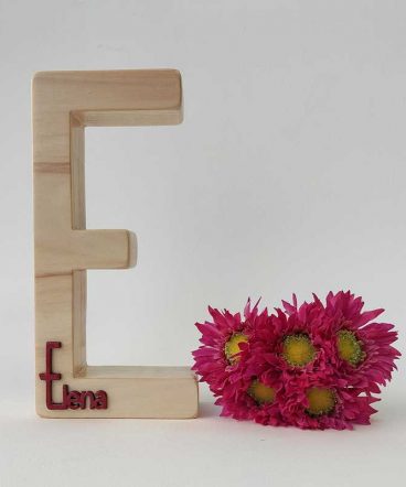 fondo blanco con letra e de madera maciza con el nombre de Elena en rojo en relieve pegado en la parte de abajo y flores fucsia a la derecha