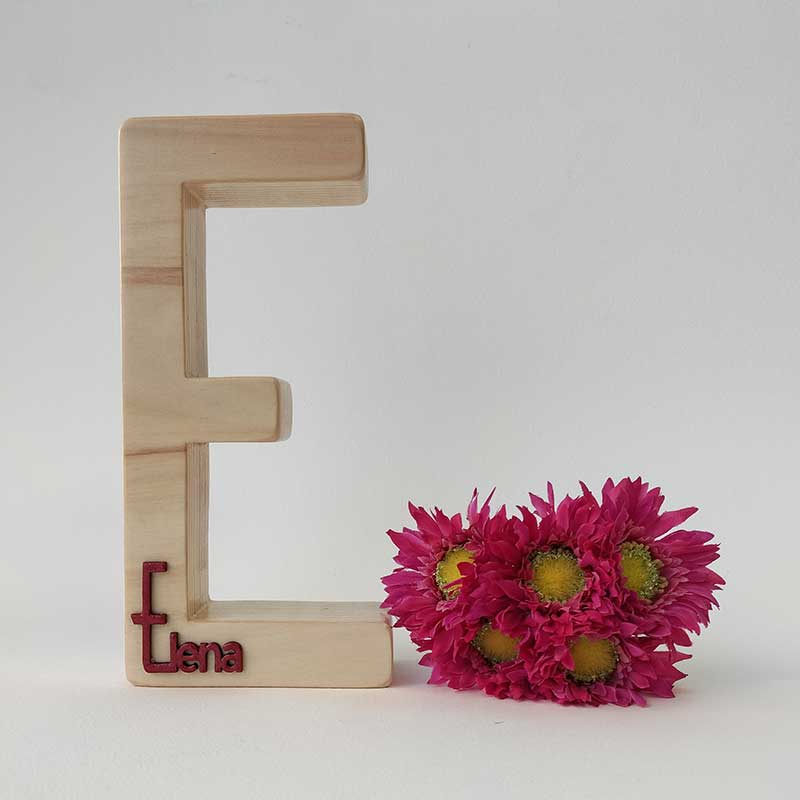 fondo blanco con letra e de madera maciza con el nombre de Elena en rojo en relieve pegado en la parte de abajo y flores fucsia a la derecha