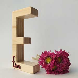 letra e de madera maciza con el nombre de Elena en rojo en relieve en la parte de abajo sobre fondo blanco y flores fucsia a la derecha