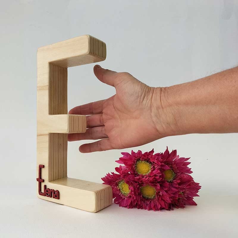fondo blanco con letra e de madera maciza con el nombre de Elena en rojo en relieve pegado en la parte de abajo, flores fucsia a la derecha y palma de una mano junto a la letra