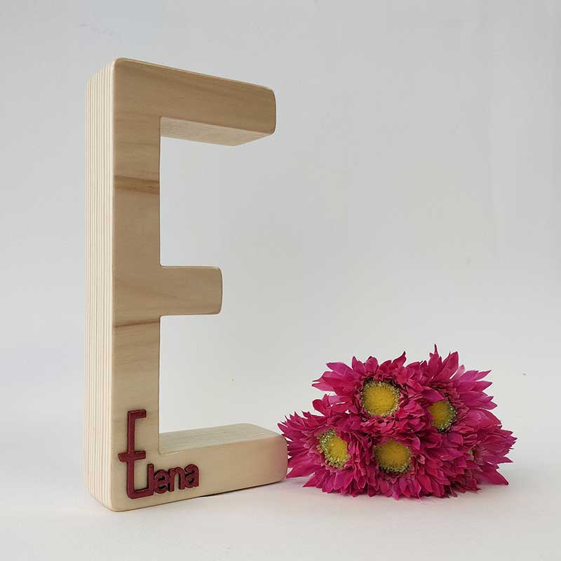 fondo blanco con letra e de madera maciza con el nombre de Elena en rojo en relieve pegado en la parte de abajo y flores fucsia a la derecha