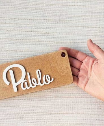 fondo con rayas suave y mano sujetando tablero de madera rectangular con el nombre de Pablo en blanco y en relieve pegado en el centro