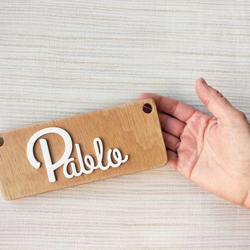 fondo con rayas suave y mano sujetando tablero de madera rectangular con el nombre de Pablo en blanco y en relieve pegado en el centro