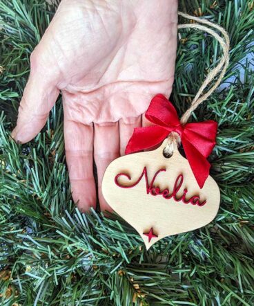 mano sujetando lágrima de navidad de madera con nombre y moña rojos