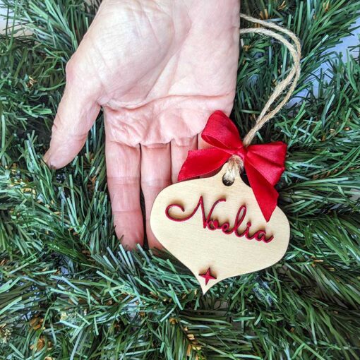 mano sujetando lágrima de navidad de madera con nombre y moña rojos