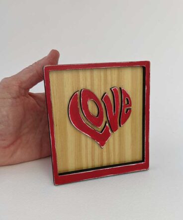 mano con cuadrito de madera con marco y la palabra LOVE en rojo y en relieve con forma de corazón