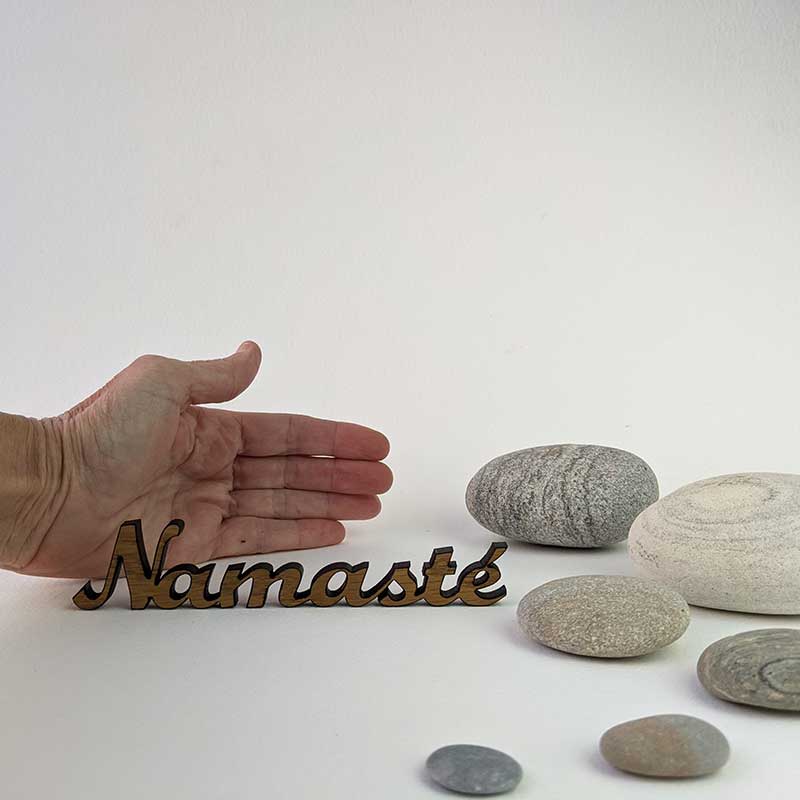 palabra namasté de roble sobre fondo blanco con piedras redondeadas y grises y una mano para saber tamaño