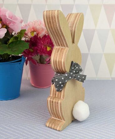 fondo grisáceo con macetas y delante silueta de conejo de pascua de madera con pompón blanco y lazo gris en el cuello