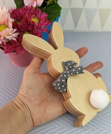 fondo gris con mano sujetando silueta de conejo de pascua de madera con lazo gris en el cuello y pompon blanco pegado