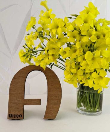 fondo blanco con vaso con flores amarillas y letra A de madera de roble con nombre Anna grabado