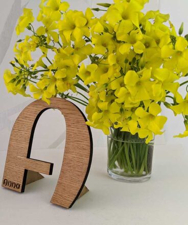 fondo blanco con vaso con flores amarillas y letra A de madera de roble con nombre Anna grabado