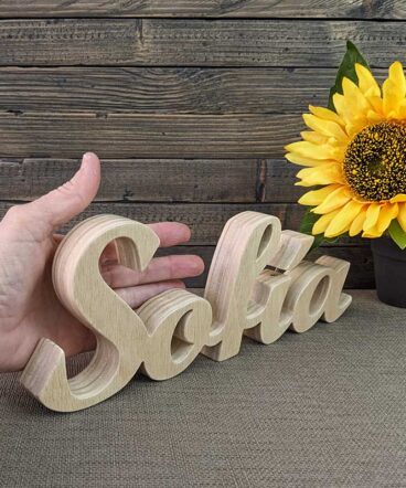nombre Sofía hecho en madera natural sobre fondo de tablas oscuras y flor amarilla al fondo