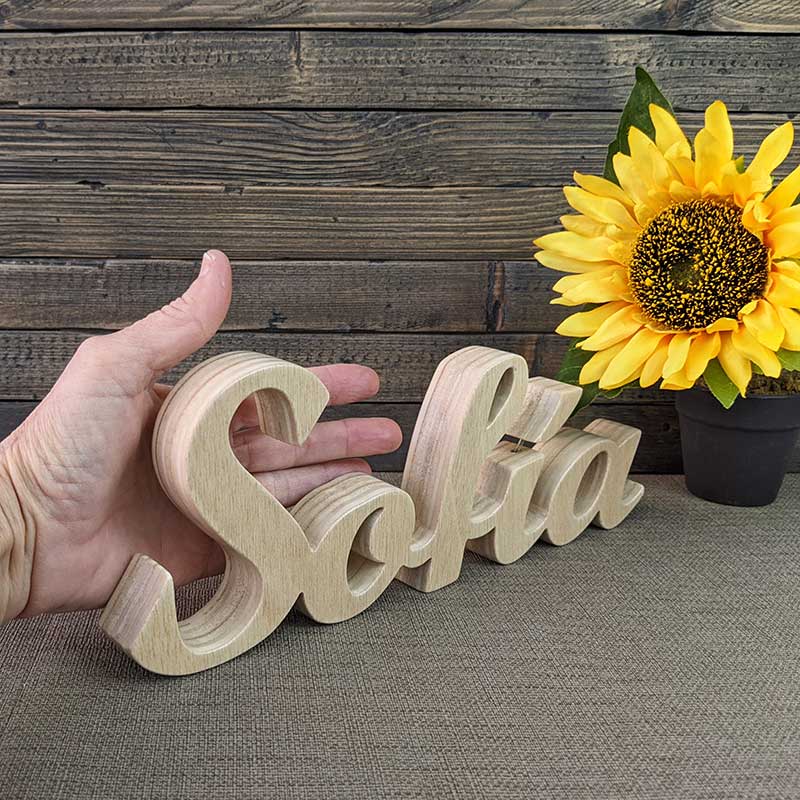 nombre Sofía hecho en madera natural sobre fondo de tablas oscuras y flor amarilla al fondo