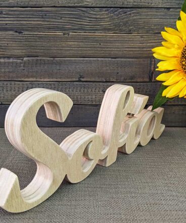 nombre Sofía hecho en madera natural sobre fondo de tablas oscuras y flor amarilla al fondo
