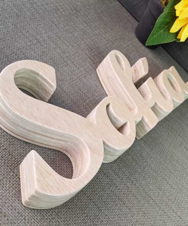 detalle del nombre de Sofía hecho en madera natural sobre superficie gris y flor amarilla al fondo