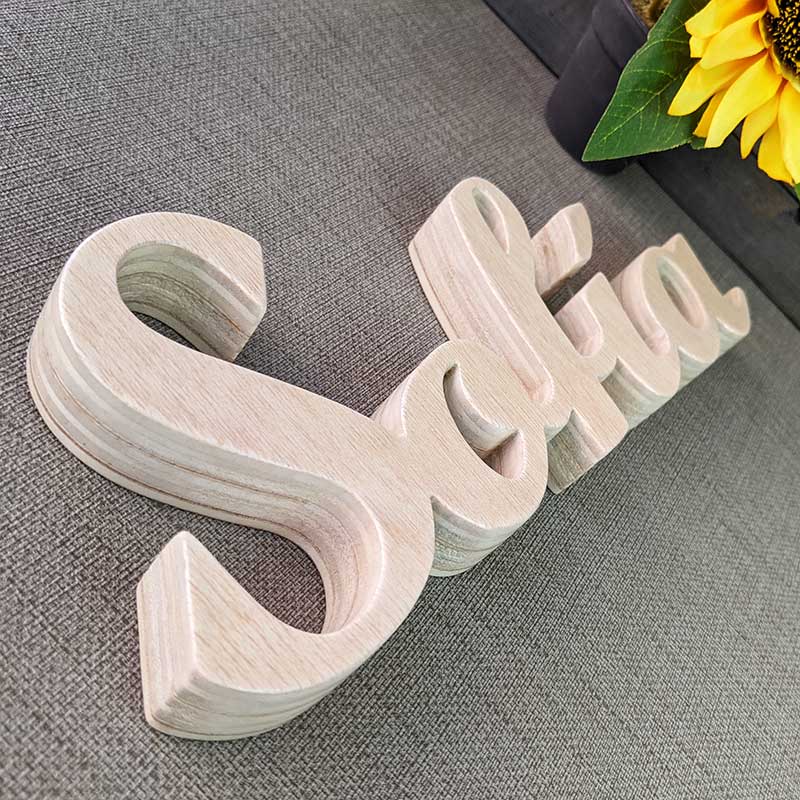 detalle del nombre de Sofía hecho en madera natural sobre superficie gris y flor amarilla al fondo