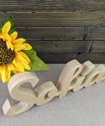 nombre Sofía hecho en madera natural sobre fondo de tablas oscuras y flor amarilla al fondo